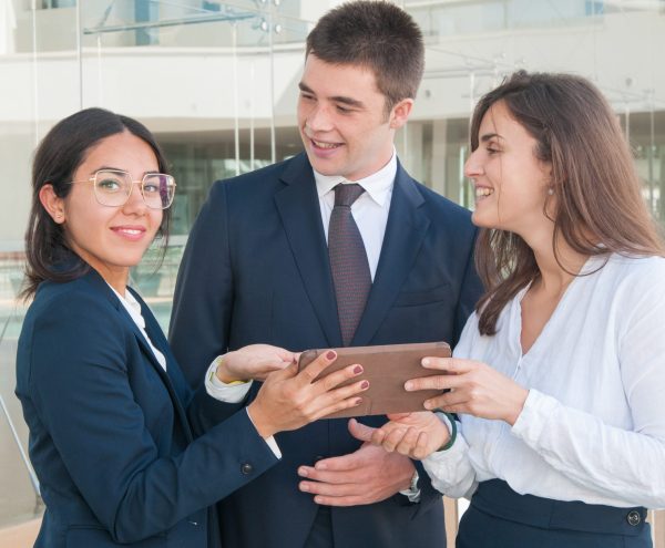 Attractive woman in spectacles showing colleagues data on tablet, looking at camera, man and woman looking with enthusiasm at her. Work, communication concept