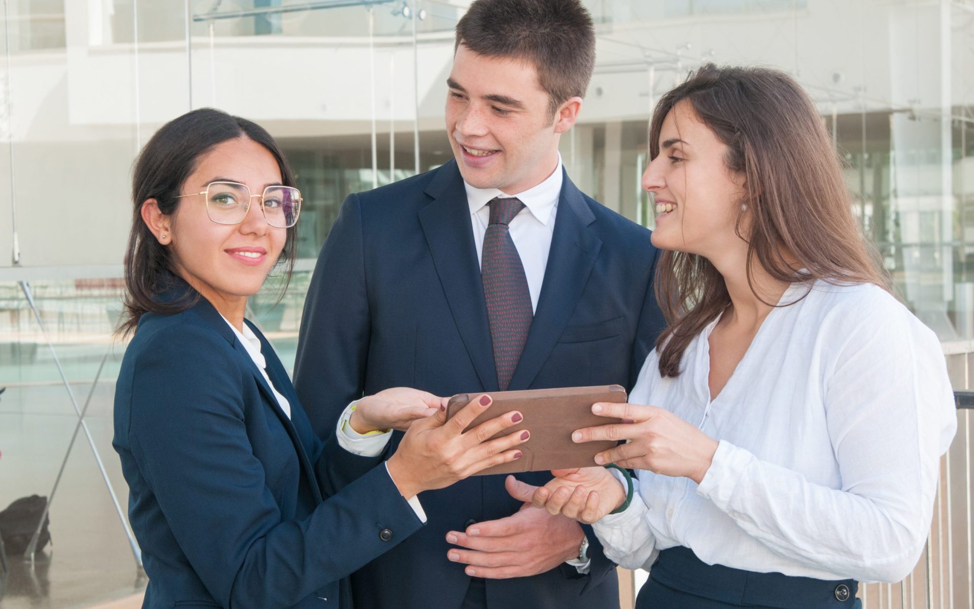 Attractive woman in spectacles showing colleagues data on tablet, looking at camera, man and woman looking with enthusiasm at her. Work, communication concept